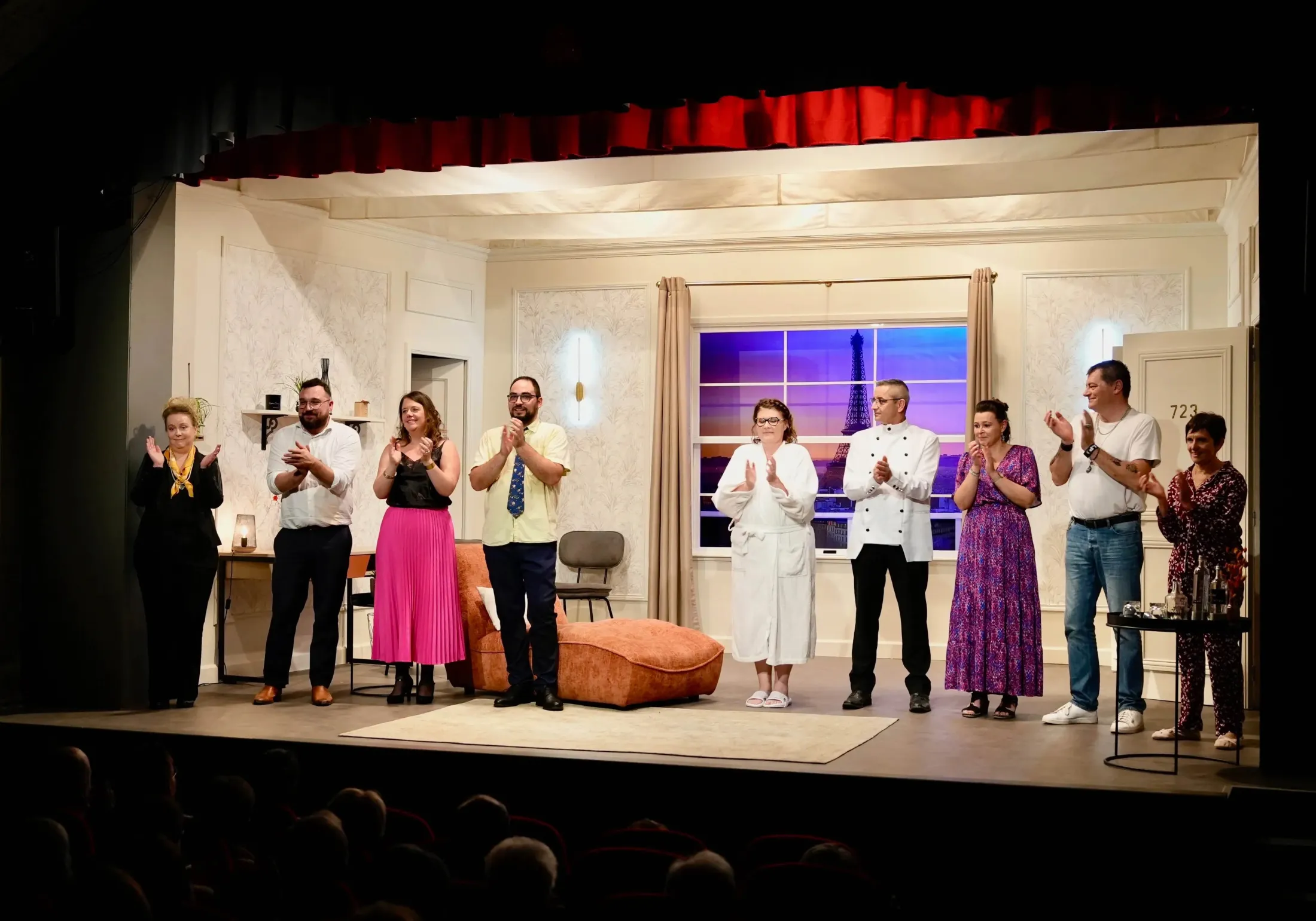 Neuf comédiens de la troupe applaudissent le public depuis la scène, dans un décor de chambre d'hôtel avec vue sur la tour Eiffel
