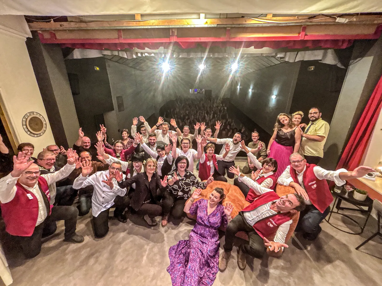 Les bénévoles de la troupe, en gilet rouge ou en costume, saluent les bras levés depuis la scène après la dernière de 2025, la salle comble en arrière-plan